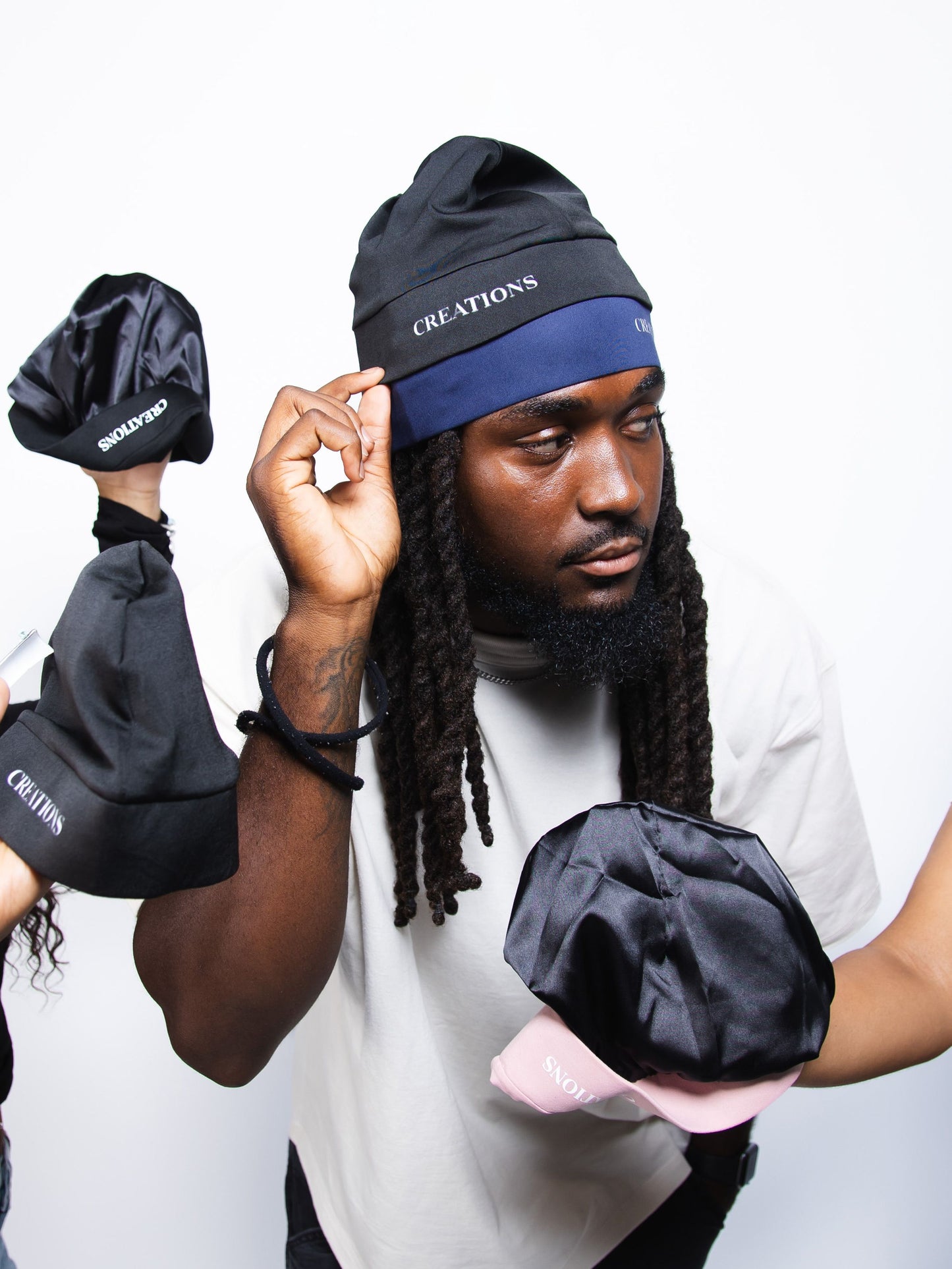Person wearing a blue beanie with 'CREATIONS' branding, holding black hats against a white background